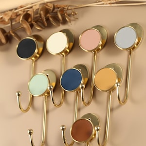 Könnte beinhalten: Ein Set aus acht dekorativen Wandhaken mit goldener Oberfläche. Jeder Haken hat einen kreisförmigen Akzent in verschiedenen Farben, darunter Schwarz, Weiß, Rosa, Grau, Grün, Blau, Orange und Braun. Die Haken sind zum Aufhängen von Gegenständen gedacht.