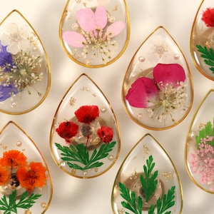 May include: Close-up of several teardrop-shaped resin pendants with gold edges, each containing dried flowers in various colors like pink, red, orange, and purple. Some pendants have small pearls and gold flecks.