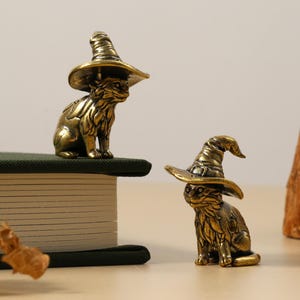 May include: Two brass cat figurines wearing witch hats. One cat sits atop a green book, while the other sits on a light-colored surface. The figurines have intricate details and a metallic finish.