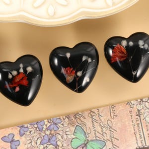 May include: Three heart-shaped decorative objects with a glossy black finish. Each heart features a dried red flower and white accents. The hearts are arranged on a light brown surface, with a decorative plate in the background.