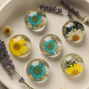 May include: Six clear resin spheres, each containing a flower in yellow, blue, or white, and gold flakes. The spheres are displayed in a white bowl, with lavender sprigs and small white flowers. The spheres are approximately 3 cm in diameter.