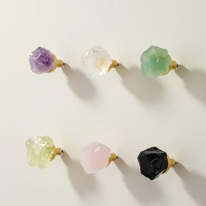 May include: Six decorative cabinet knobs with a natural, raw crystal appearance. The knobs are in various colors, including purple, clear, green, yellow, pink, and black. Each crystal is mounted on a gold-colored base.