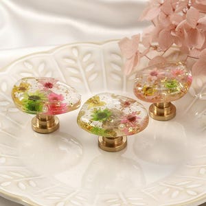 Könnte beinhalten: Drei dekorative Schrankknöpfe mit Blumeneinlagen. Die Knöpfe haben eine transparente Harzoberseite mit eingebetteten rosa, gelben und grünen Blumen. Sie sind auf messingfarbenen Sockeln montiert und liegen auf einem weißen, gewellten Teller.