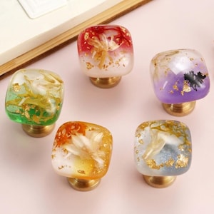 Puede incluir: Cinco tiradores decorativos para armarios con tapas cuadradas de resina en varios colores, incluyendo verde, naranja, rojo, morado y transparente. Cada tirador presenta detalles dorados y una base de latón. Los tiradores están dispuestos sobre una superficie rosa.