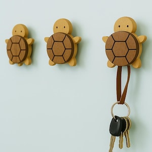 Könnte beinhalten: Drei hölzerne Wandhaken in Schildkrötenform. Die Schildkröten haben hellbraune Körper und dunkelbraune Schalenmuster. Eine Schildkröte hält einen Lederriemen mit einem Schlüsselring und Schlüsseln. Die Haken sind an einer hellblauen Wand befestigt.