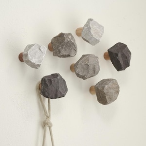 Könnte beinhalten: Ein Set aus sieben dekorativen Wandhaken. Jeder Haken hat ein Naturstein-Design in Grau- und Brauntönen, montiert auf einem Holzsockel. Ein beiges Seil hängt an einem der Haken. Die Haken sind an einer weißen Wand befestigt.