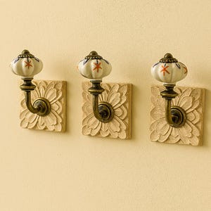 Könnte beinhalten: Drei dekorative Wandhaken. Jeder Haken hat eine cremefarbene, floral gemusterte quadratische Basis mit einem bronzefarbenen Haken. Der Haken trägt einen weißen Keramikknauf mit roten Sternenmustern und blauen Akzenten.