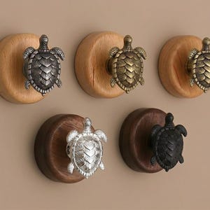 May include: Five decorative turtle-shaped knobs in various metallic finishes, mounted on wooden discs. The knobs include silver, bronze, and black options, offering a nautical or coastal theme for furniture or decor.
