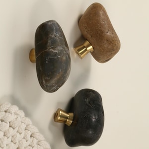 May include: Three natural stone cabinet knobs with brass hardware. The knobs are in shades of brown, gray, and black. The stone has a smooth, rounded shape. The knobs are mounted on a white surface.