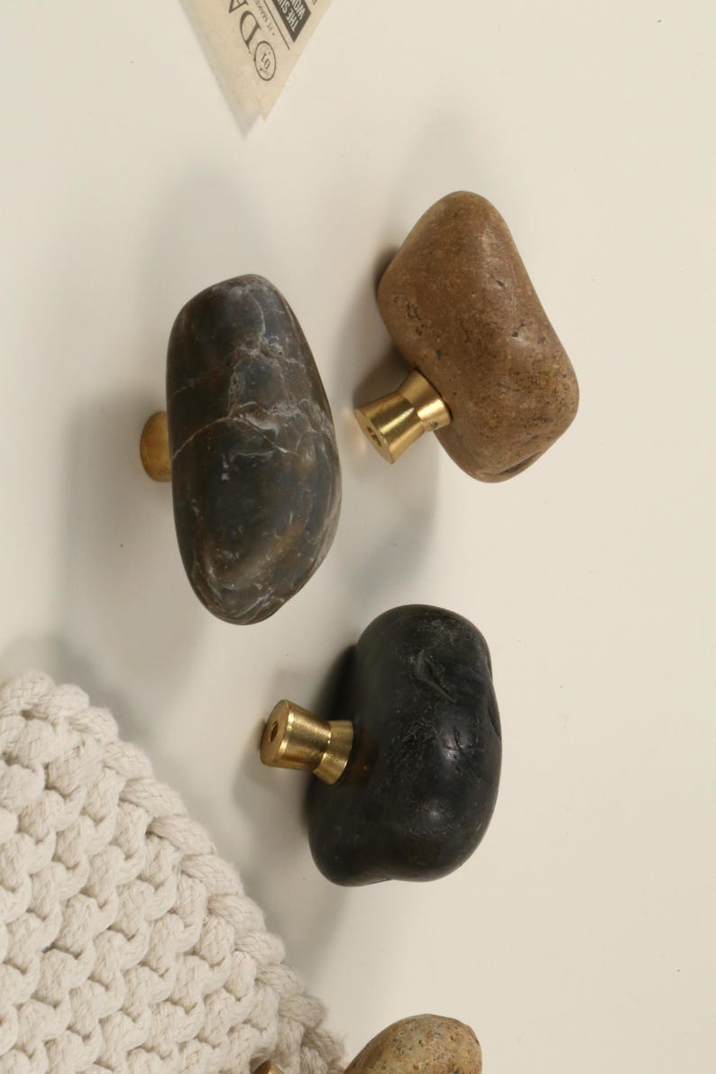 May include: Three natural stone cabinet knobs with brass hardware. The knobs are in shades of brown, gray, and black. The stone has a smooth, rounded shape. The knobs are mounted on a white surface.