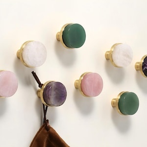 以下が含まれることがあります： 真鍮ベースとカラフルな石のトップが付いた、8つの装飾的なキャビネットノブのコレクション。ノブはピンク、緑、白、紫など、さまざまな色があります。茶色のバッグがノブの1つから吊り下げられています。