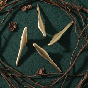 Könnte beinhalten: Fünf goldfarbene Schrankgriffe auf einer dunkelgrünen Oberfläche, umgeben von Zweigen und kleinen, braunen, strukturierten Objekten. Die Griffe haben ein glattes, längliches Design mit einer leichten Krümmung. Die Gesamtästhetik ist modern und minimalistisch.