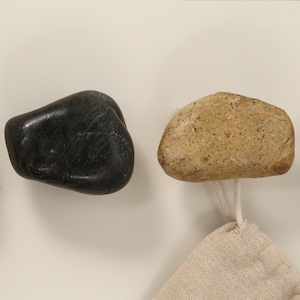 May include: A collection of natural stone hooks in various colors, including black and tan. Each hook is attached to a small, beige fabric loop, suitable for hanging items. The hooks are mounted on a white surface.