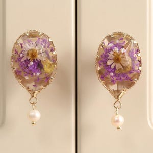 May include: Two oval-shaped cabinet knobs featuring clear resin encasing purple and white dried flowers. Each knob is framed in gold-toned metal with a wire-wrapped base and a single dangling white pearl bead attached to the bottom of the fixture.