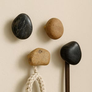 May include: Four stone-shaped wall hooks in varying shades of black and tan. One hook has a white rope attached. The hooks are mounted on a neutral-coloured wall, offering a natural, organic aesthetic.