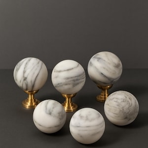 Peut inclure: Six sphères décoratives en marbre veiné de gris, certaines montées sur de courts socles en métal doré. Les sphères sont disposées sur une surface gris foncé, créant une présentation sophistiquée. Le marbre est poli.