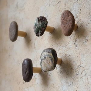 Może przedstawiać: Cztery dekoracyjne haczyki ścienne wykonane z naturalnych kamieni rzecznych i drewnianych kołków. Kamienie mają różne kolory, w tym odcienie brązu, szarości i zieleni. Haczyki są zamontowane na teksturowanej, jasnej ścianie.