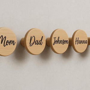 May include: A set of five wooden wall hooks with a natural finish. Each hook has a circular design with black script text that reads "Mom", "Dad", "Johnson", "Hanna", and "Hanna".