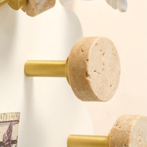 May include: Close-up of decorative cabinet knobs. Each knob features a cylindrical, beige stone top attached to a gold-colored metal stem. The knobs are mounted on a white surface, suggesting a furniture or decor theme.