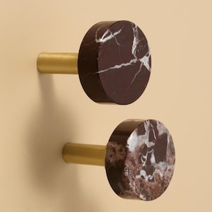 May include: Two cabinet knobs. Each knob has a circular, dark red marble face with white veining, mounted on a gold-coloured cylindrical stem. The knobs are set against a neutral, light beige background.