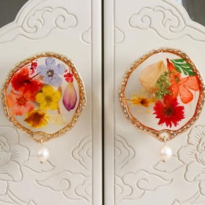 May include: Two circular cabinet knobs featuring clear resin centers embedded with colorful pressed flowers, including red, yellow, and blue petals. Each knob has a textured gold-tone metal rim and a small white pearl-like bead hanging from the bottom.