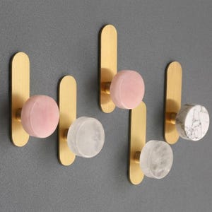 May include: A set of five decorative wall hooks. Each hook features a gold-colored, elongated backplate with a round knob. The knobs are made of pink, clear, and white marble-like materials. The hooks are mounted on a gray wall.