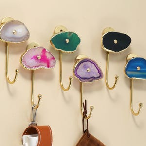 May include: A set of six decorative wall hooks with gold-colored metal hardware. Each hook features a unique, colorful agate stone slice with gold edging. The hooks are mounted on a neutral-colored wall, with a leather item hanging from one.