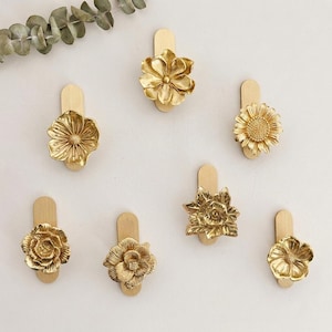 Può includere: Una collezione di sette ganci da parete dorati a forma di fiore. Ogni gancio presenta un diverso design floreale, tra cui girasoli e rose, montato su una piastra posteriore rettangolare. I ganci sono disposti su uno sfondo bianco.