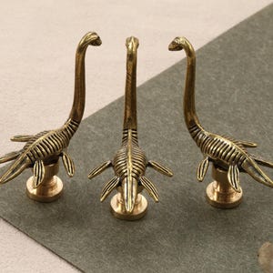 May include: Three bronze-colored dinosaur-shaped wax seal stamps. Each stamp features a long-necked, aquatic dinosaur design with detailed ribbing and flipper-like appendages, mounted on a round base.