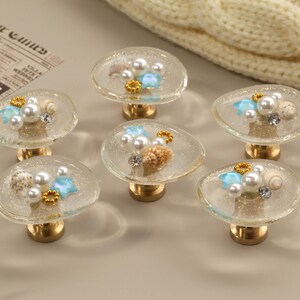 May include: Six decorative cabinet knobs with a coastal theme. Each knob features a clear, oval-shaped resin top with embedded seashells, pearls, and gold accents. The knobs have a gold-coloured base.