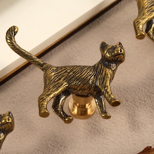 Könnte beinhalten: Schrankknöpfe in Form einer Katze aus Bronze. Die Knöpfe sind mit strukturiertem Fell und einer Messingoberfläche versehen. Die Katze ist in einer Laufpose mit nach oben gebogenem Schwanz dargestellt. Diese dekorativen Knöpfe sind für Möbel geeignet.
