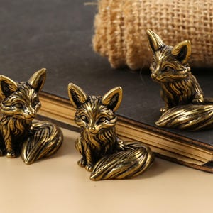 May include: Three bronze fox figurines in various poses. The foxes have detailed fur and are sitting or reclining. The figurines are on a dark surface with a burlap texture in the background. The foxes are a decorative item.