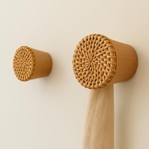 Op de afbeelding: Twee houten wandhaken met gevlochten rotan gezichten. Eén haak houdt een beige stof vast. De haken zijn cilindrisch met een natuurlijke houten afwerking. Het gevlochten rotan voegt een decoratief tintje toe.