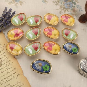 Op de afbeelding: Een verzameling ovale hars cabochons met goudkleurige randen. Elke cabochon bevat gedroogde bloemen in roze, gele en groene tinten. Eén cabochon is blauw met witte en groene bloemen. De cabochons zijn gerangschikt op een crèmekleurig oppervlak.