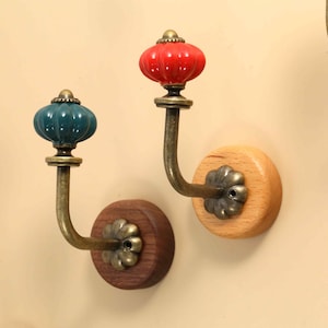 Op de afbeelding: Wandhaken met een gebogen arm van antiek messing, een ronde houten basis en een decoratieve, gegroefde keramische knop. De knoppen zijn verkrijgbaar in effen kleuren zoals rood, blauwgroen en lichtroze, bevestigd op licht of donker hout.