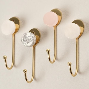 May include: Four decorative wall hooks with a gold finish. Each hook features a circular accent in white, pink, or marble-patterned stone. The hooks are designed with a curved, elongated shape, perfect for hanging items.