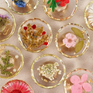 Könnte beinhalten: Eine Sammlung von transparenten Harzuntersetzern mit Goldkanten, die jeweils getrocknete Blumen in verschiedenen Farben enthalten. Einige Untersetzer zeigen rosa, weiße und gelbe Blumen, während andere rote und violette Akzente haben. Die Untersetzer sind auf einer hellen Oberfläche angeordnet.