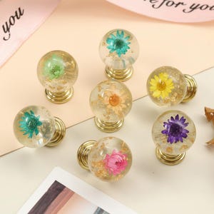May include: A collection of seven clear, spherical cabinet knobs with gold-colored bases. Each knob encases a different colored dried flower, including green, yellow, purple, pink, and orange, with gold flakes. The knobs are arranged on a light-colored surface.