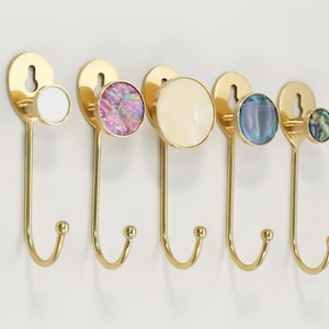 Könnte beinhalten: Eine Reihe von fünf dekorativen Wandhaken mit goldener Oberfläche. Jeder Haken hat einen anderen runden, farbenfrohen Akzent: weiß, rosa, creme, blau und grün. Die Haken sind zum Aufhängen von Gegenständen gedacht.