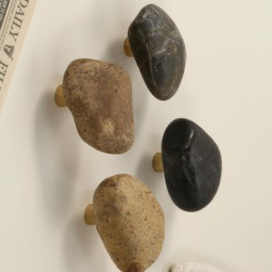 May include: Four stone cabinet knobs in varying shades of brown and black, each with a brass base. The knobs are smooth and rounded, resembling natural river stones. A newspaper clipping is visible in the background.