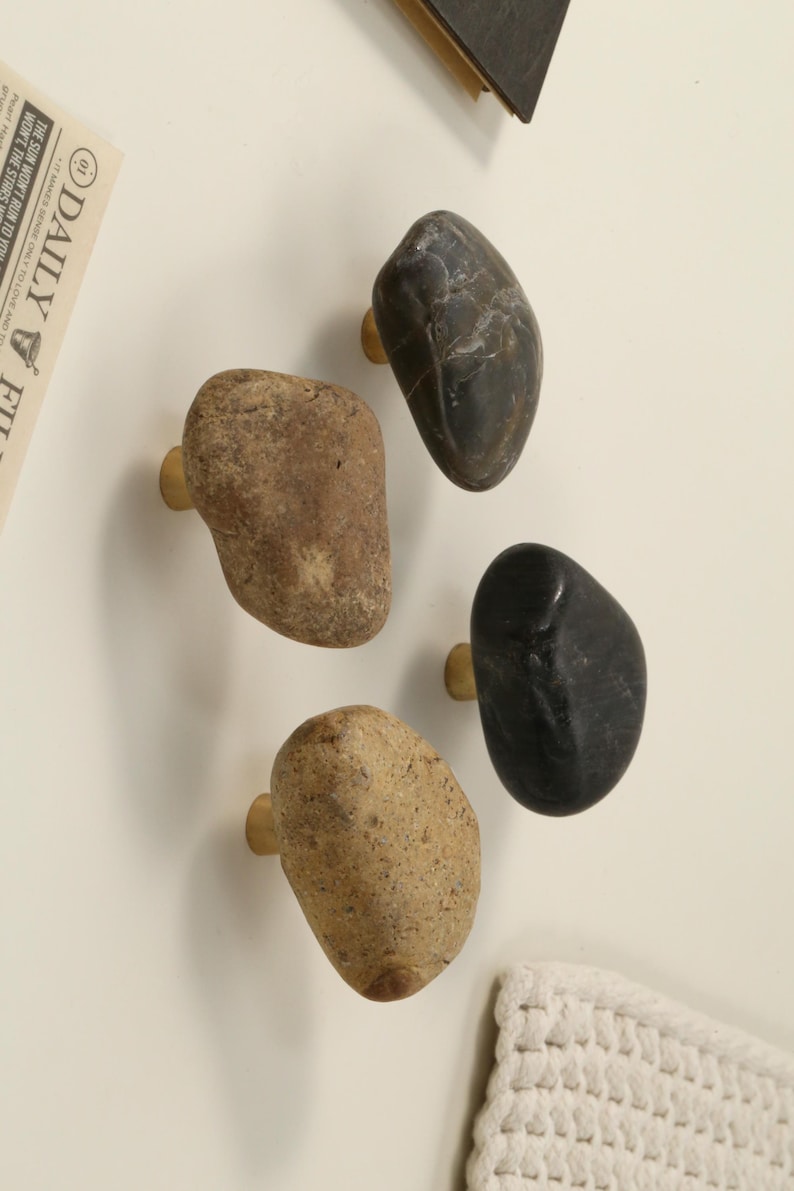 May include: Four stone cabinet knobs in varying shades of brown and black, each with a brass base. The knobs are smooth and rounded, resembling natural river stones. A newspaper clipping is visible in the background.