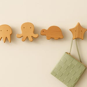 Może przedstawiać: Drewniane wieszaki ścienne w kształcie meduzy, ośmiornicy, żółwia i gwiazdy. Wieszaki wykonane są z jasnego drewna i zamontowane na kremowej ścianie. Zielony portfel w kwadratowy wzór wisi na wieszaku w kształcie gwiazdy.