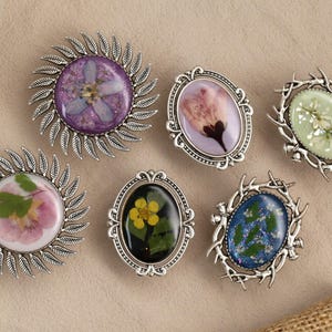 Puede incluir: Colección de seis broches hechos a mano con flores secas encapsuladas en resina. Los broches tienen marcos de metal plateado en varias formas, incluyendo diseños ovalados y circulares. Las flores son de color púrpura, rosa, amarillo y blanco.