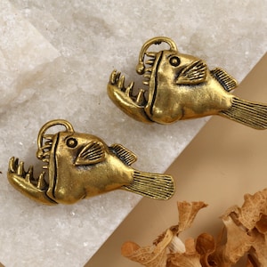 Peut inclure: Deux breloques antiques en forme de baudroie dorée. Chaque breloque présente un motif de poisson détaillé avec des mâchoires ouvertes, une nageoire dorsale et une boucle d'attache. Les breloques sont présentées sur une surface blanche et beige.