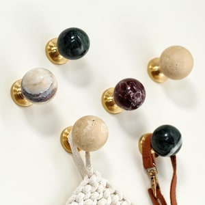 Può includere: Una collezione di pomelli decorativi per mobili con basi in ottone e sfere di marmo in vari colori, tra cui verde, beige e viola. Un pomello tiene un oggetto in macramè e un altro un cinturino in pelle.