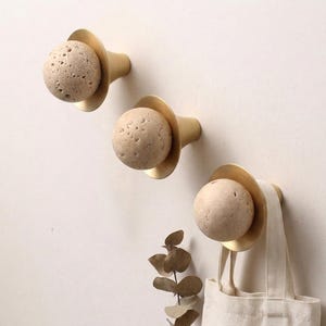 May include: Three wall hooks with a brass finish and a beige, spherical stone accent. One hook holds a beige tote bag. The hooks are mounted on a light beige wall. Decorative and functional.