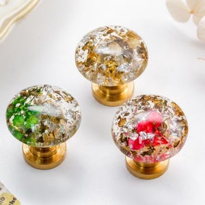 Puede incluir: Tres pomos decorativos para armarios con tapas redondas y transparentes llenas de copos de pan de oro y plata y acentos coloridos. Los pomos tienen bases de latón y están dispuestos sobre una superficie blanca.