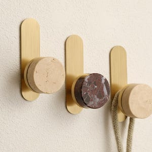 Peut inclure: Trois patères murales avec une finition en laiton et des accents en pierre cylindriques. Les crochets présentent une pierre beige clair, rouge foncé et blanche, et beige clair. Une corde beige est suspendue au troisième crochet.