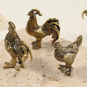 Op de afbeelding: Een verzameling antieke messing hanen- en hennenbeeldjes. De hanenbeeldjes staan in verschillende poses, met gedetailleerde veren en kam. Het hennenbeeldje staat. De beeldjes staan op een getextureerd oppervlak.