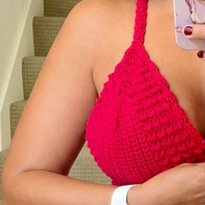 May include: A bright red, handmade crochet halter top. The top is a solid color and has a textured appearance. The straps are thick and the top is a triangle shape. A white watch with a gold case is visible on the wrist.